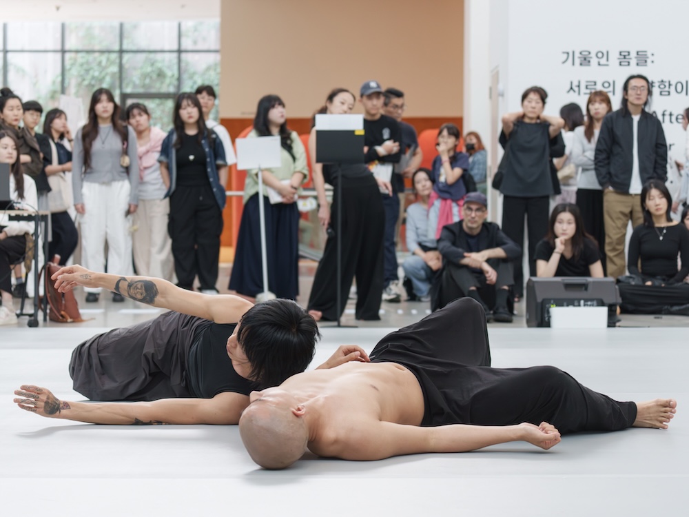 작가, 최태윤. 작품명, 내게 기대요, 부문, 벽화. 제작년도, 2025년. 벽에 실크스크린 설치, 20 ✕ 6 m. 국립현대미술관 제작지원. 두 사람이 등을 기댄 모습이 검은 선으로 그려져 있다. ‘내게 기대요’라는 문구가 한쪽에 쓰여 있다. 평화로운 표정이다.