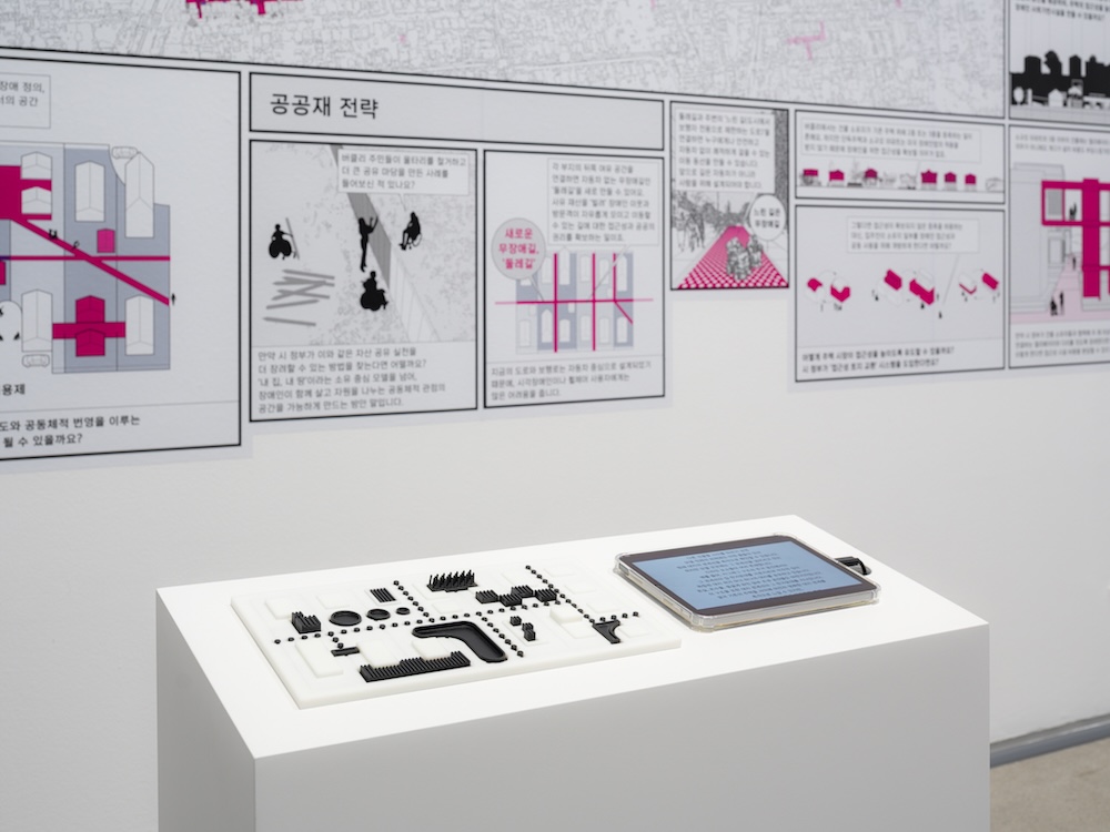 작가, 데이비드 기슨, 브렛 스나이더, 아이린 챙. 작품명, 블록 파티: 자립생활에서 장애 공동체에 이르기까지. 부분, 설치. 제작년도, 2022년 (2025년 재제작), 매체 및 기법, 모형, 그래픽, 소리. 규격, 가변 크기. 어느 마을을 본따 만든 모형이 놓여 있다. 모형 가운데에는 공용 수영장이 있고, 분홍색으로 공유지가 표시되어 있다. 뒤쪽에는 벽면을 활용하여 만화가 그려져 있다.
