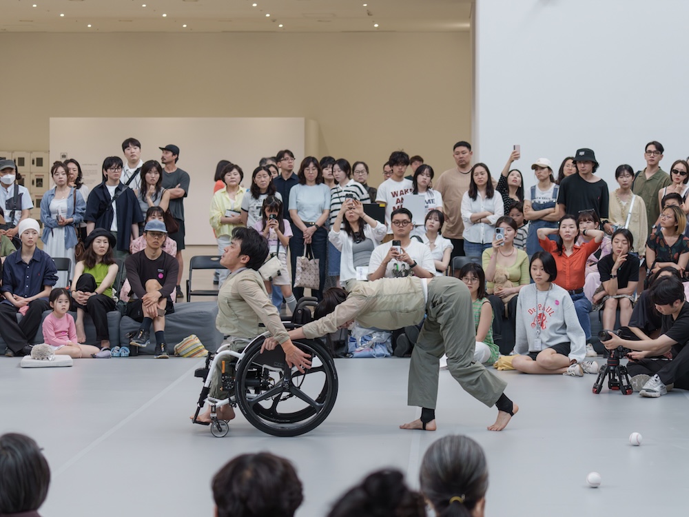작가, 김원영. 작품명, 보철물로서 움직이기, 머신, 어포던스, 케어. 부문, 퍼포먼스. 제작년도, 2025년. 국립현대미술관 제작지원. 영상 스틸. 두 사람이 춤을 추고 있다. 한 사람은 휠체어를 타고 있다. 두 사람은 두 손을 바닥에 짚고 네 발로 움직이고 있다. 하나의 축을 중심으로 원형을 그리면서 돌고 있다.