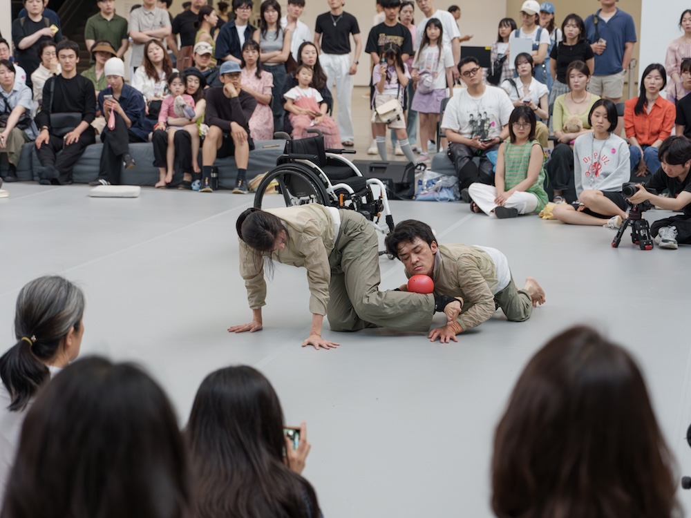 작가, 김원영. 작품명, 보철물로서 움직이기, 머신, 어포던스, 케어. 부문, 퍼포먼스. 제작년도, 2025년. 국립현대미술관 제작지원. 영상 스틸. 두 사람이 춤을 추고 있다. 한 사람은 휠체어를 타고 있다. 두 사람은 두 손을 바닥에 짚고 네 발로 움직이고 있다. 하나의 축을 중심으로 원형을 그리면서 돌고 있다.