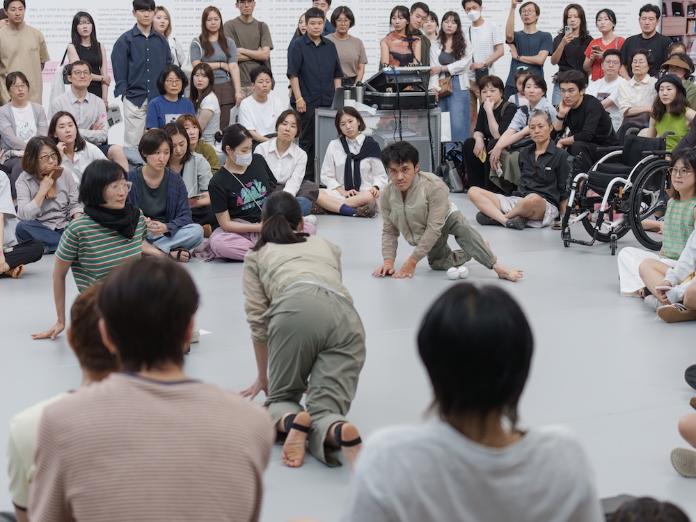 작가, 김원영. 작품명, 보철물로서 움직이기, 머신, 어포던스, 케어. 부문, 퍼포먼스. 제작년도, 2025년. 국립현대미술관 제작지원. 영상 스틸. 두 사람이 춤을 추고 있다. 한 사람은 휠체어를 타고 있다. 두 사람은 두 손을 바닥에 짚고 네 발로 움직이고 있다. 하나의 축을 중심으로 원형을 그리면서 돌고 있다.