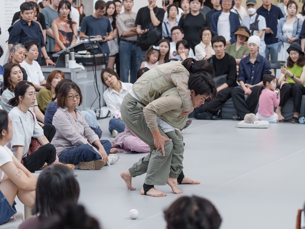 작가, 김원영. 작품명, 보철물로서 움직이기, 머신, 어포던스, 케어. 부문, 퍼포먼스. 제작년도, 2025년. 국립현대미술관 제작지원. 영상 스틸. 두 사람이 춤을 추고 있다. 한 사람은 휠체어를 타고 있다. 두 사람은 두 손을 바닥에 짚고 네 발로 움직이고 있다. 하나의 축을 중심으로 원형을 그리면서 돌고 있다.