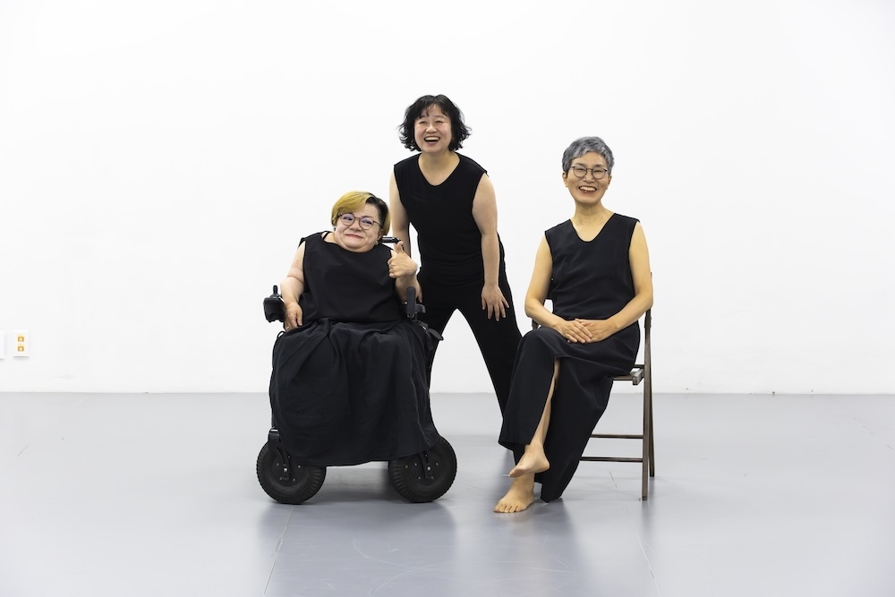 작가, 김영옥, 조미경, 이진희. 작품명, 파도 나이듦과 장애의 교차적 만남. 부문, 퍼포먼스. 제작년도, 2025년. 국립현대미술관 제작지원. 나이 든 몸과 장애가 있는 몸이 함께 등장하는 공연을 위한 홍보 사진이다. 두 사람이 미술관 앞 마당에 서 있다. 한 사람은 휠체어를 탔다. 한 사람은 백발이다. 둘 모두 검은 드레스를 입었다.