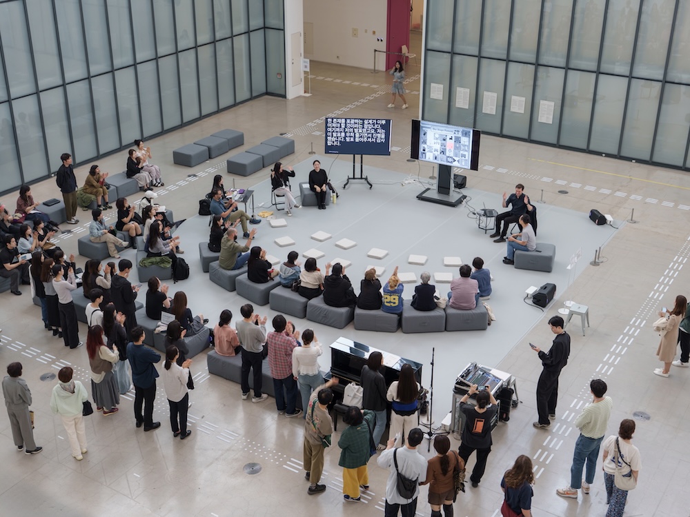 작가, 리처드 도허티. 작품명, 농인공간: 입을 맞추는 의자. 부문, 설치. 제작년도, 2025년. 매체 및 기법, 철에 도장, 가변 설치. INTG, 국립서울농학교, 다니엘 모레노 제작협력. 국립현대미술관 제작지원. 미술관 정문 계단 위에 노란색, 하늘색, 주황색 벤치가 놓여 있다. 사람들이 서로 마주보고 앉을 수 있도록 방향 지워져 있다. 벤치는 계단을 통로가 아닌 휴식공간으로 바꾼다. 미술관에 들어가려는 사람들은 모두 측면의 경사로를 이용하게 된다.