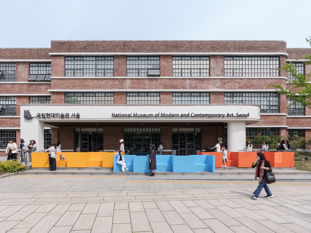 작가, 리처드 도허티. 작품명, 농인공간: 입을 맞추는 의자. 부문, 설치. 제작년도, 2025년. 매체 및 기법, 철에 도장, 가변 설치. INTG, 국립서울농학교, 다니엘 모레노 제작협력. 국립현대미술관 제작지원. 미술관 정문 계단 위에 노란색, 하늘색, 주황색 벤치가 놓여 있다. 사람들이 서로 마주보고 앉을 수 있도록 방향 지워져 있다. 벤치는 계단을 통로가 아닌 휴식공간으로 바꾼다. 미술관에 들어가려는 사람들은 모두 측면의 경사로를 이용하게 된다.