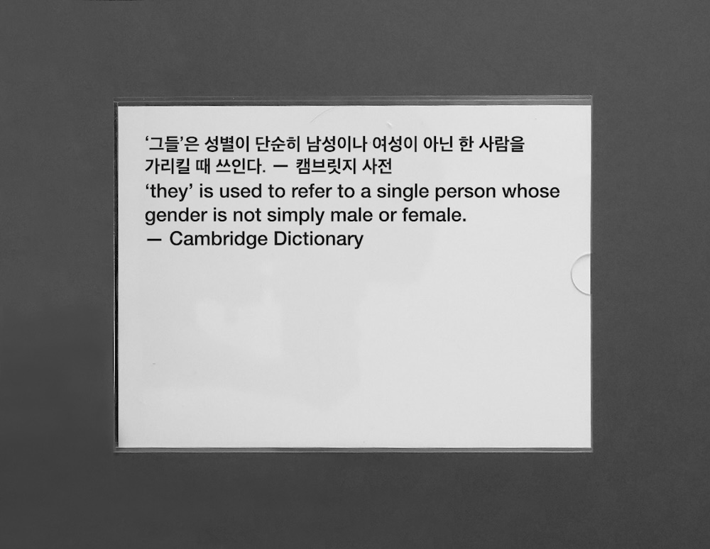 작가, 윤충근. 작품명, 격언. 부문, 설치. 제작년도, 2025년. 매체 및 기법, 아크릴에 인쇄, 가변 설치. 국립현대미술관 제작지원. 투명한 아크릴 판 안에 격언이 쓰여 있다.여느 화장실에서 볼 수 있는 것과 같은 평범한 디자인이다.  “Ey/em/eir(s)은 they/them/theirs를 줄여 말하는 성중립 대명사이다.”