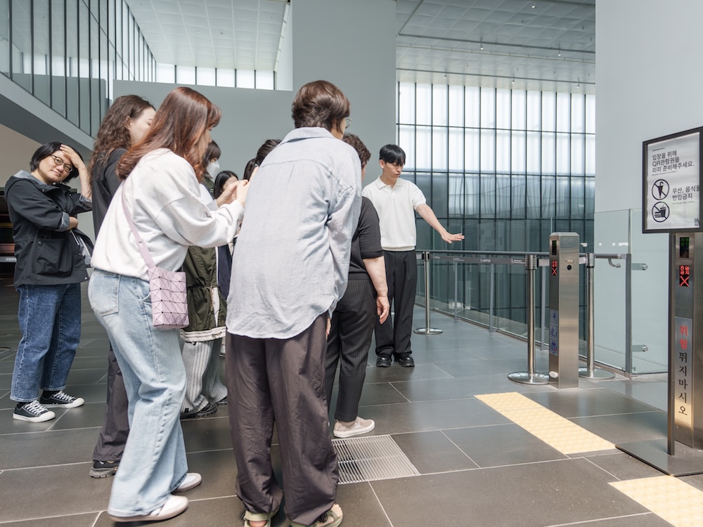 작가, 윤충근. 작품명, 격언. 부문, 설치. 제작년도, 2025년. 매체 및 기법, 아크릴에 인쇄, 가변 설치. 국립현대미술관 제작지원. 투명한 아크릴 판 안에 격언이 쓰여 있다.여느 화장실에서 볼 수 있는 것과 같은 평범한 디자인이다.  “Ey/em/eir(s)은 they/them/theirs를 줄여 말하는 성중립 대명사이다.”